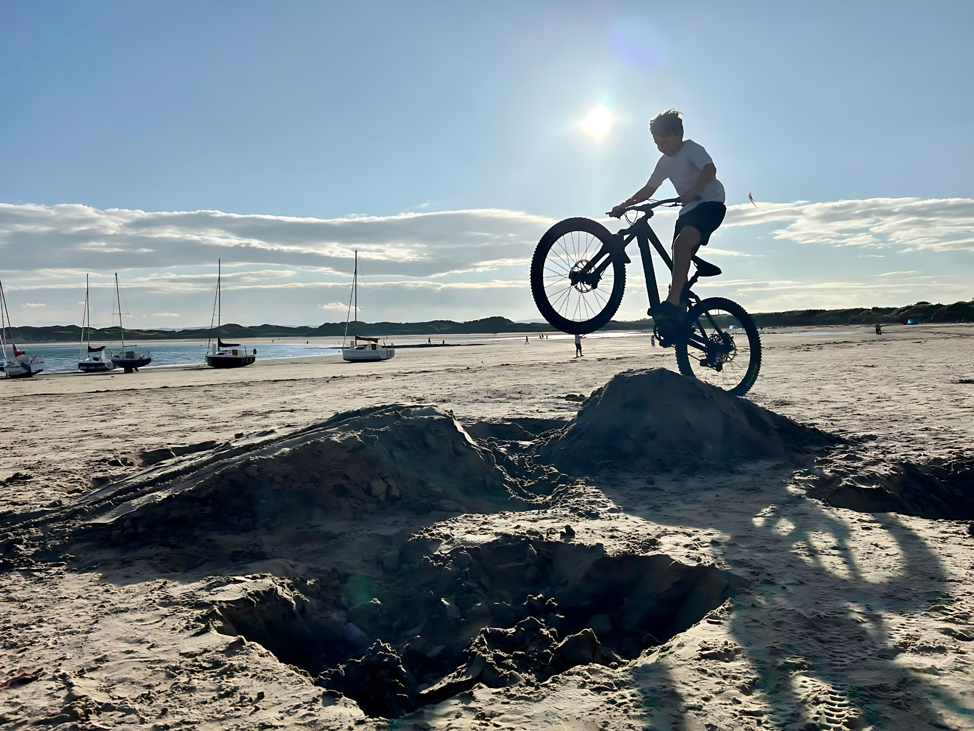 Beadnell Bay Biking A cyclist performing a stunt on a beach, with boats in the background and sunlight shining.