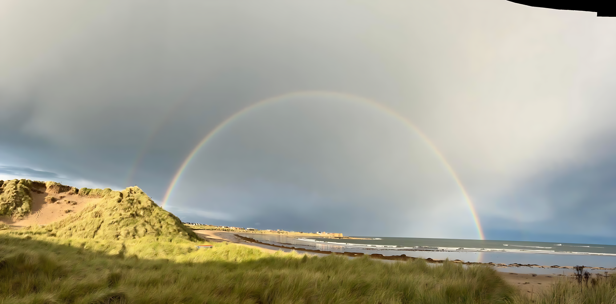 Beadnell Bay Rainbow Rainbow arching over a grassy landscape and calm waters beneath a cloudy sky.