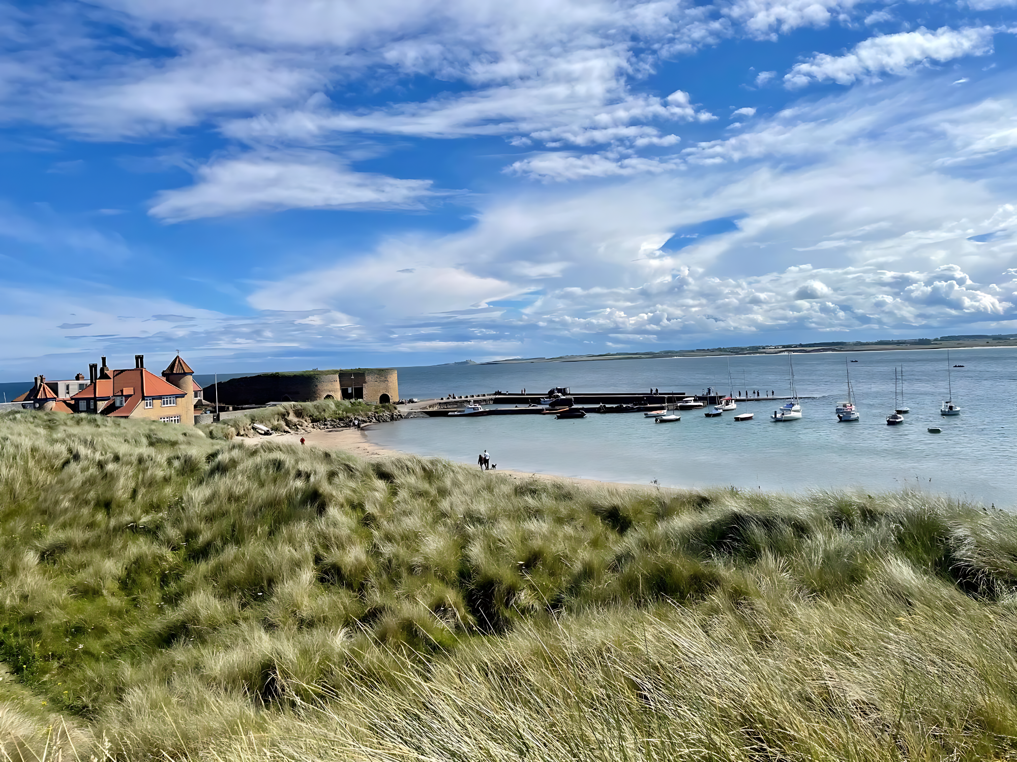 Beadnell Bay and Harbour Seaside view with boats, shoreline, green grass, and blue sky with clouds.